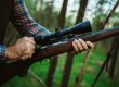 A close-up of a hunter wearing a plaid shirt holding a rifle with a wooden stock and a large optical scope in a forest setting.