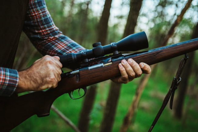 A close-up of a hunter wearing a plaid shirt holding a rifle with a wooden stock and a large optical scope in a forest setting.