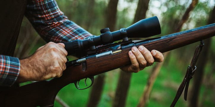 A close-up of a hunter wearing a plaid shirt holding a rifle with a wooden stock and a large optical scope in a forest setting.
