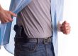 A man lifts his unbuttoned blue shirt to reveal a black handgun holstered inside the waistband of his jeans, demonstrating a concealed carry setup.