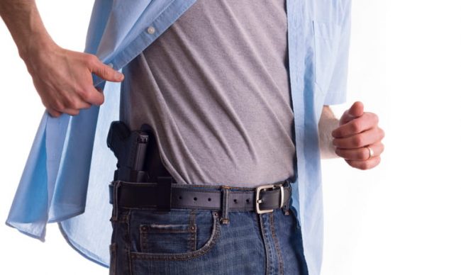 A man lifts his unbuttoned blue shirt to reveal a black handgun holstered inside the waistband of his jeans, demonstrating a concealed carry setup.
