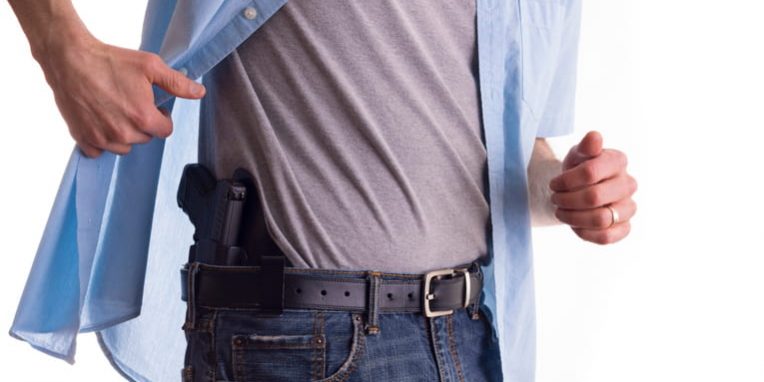 A man lifts his unbuttoned blue shirt to reveal a black handgun holstered inside the waistband of his jeans, demonstrating a concealed carry setup.