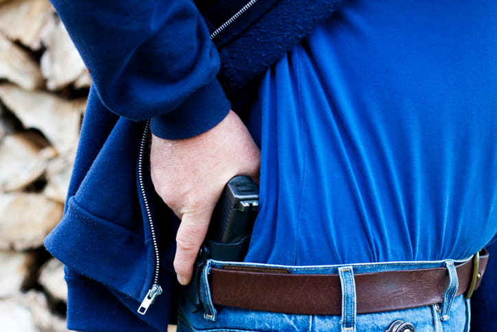 concealed-carry-under-hoodie A close-up of a person wearing a blue t-shirt and a dark blue zip-up hoodie, carrying a black handgun holstered inside the waistband of their blue jeans, secured by a brown leather belt.