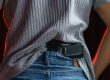 A close-up view from behind of a person tucking a black semi-automatic handgun into the back waistband of their blue jeans, underneath a gray ribbed shirt.