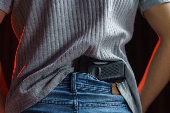 handgun-tucked-in-back-of-jeans A close-up view from behind of a person tucking a black semi-automatic handgun into the back waistband of their blue jeans, underneath a gray ribbed shirt.