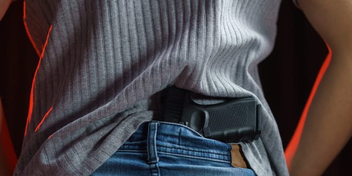 handgun-tucked-in-back-of-jeans A close-up view from behind of a person tucking a black semi-automatic handgun into the back waistband of their blue jeans, underneath a gray ribbed shirt.
