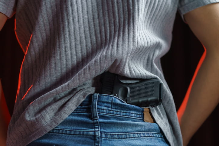 handgun-tucked-in-back-of-jeans A close-up view from behind of a person tucking a black semi-automatic handgun into the back waistband of their blue jeans, underneath a gray ribbed shirt.