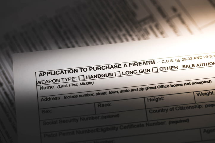 A close-up, top-down view of an official "Application to Purchase a Firearm" document with fields for weapon type, name, address, and personal descriptors