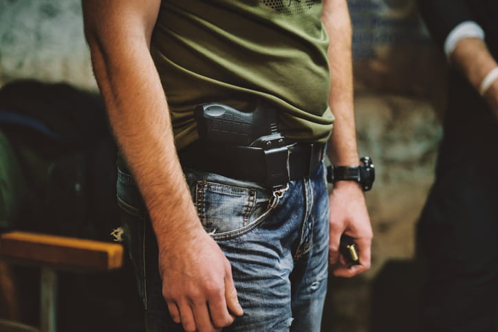 Close-up of a man wearing a handgun in a concealed carry holster attached to his belt, tucked under a green t-shirt with casual denim jeans.
