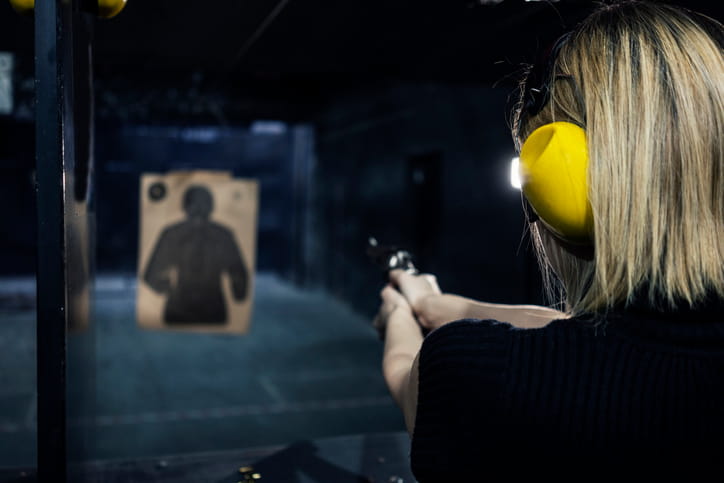 Over-the-shoulder view of a woman wearing yellow ear protection aiming a revolver at a paper silhouette target in an indoor shooting range.