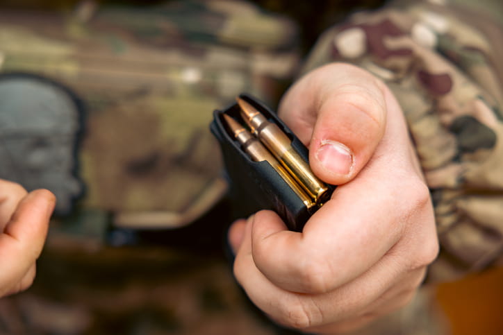 A close-up shot of a person’s hand holding a black rifle magazine loaded with several brass-cased bullets. The person is wearing a camouflage jacket in the background. The focus is sharp on the metal cartridges and the magazine's top edge, showing the ammunition ready for use.