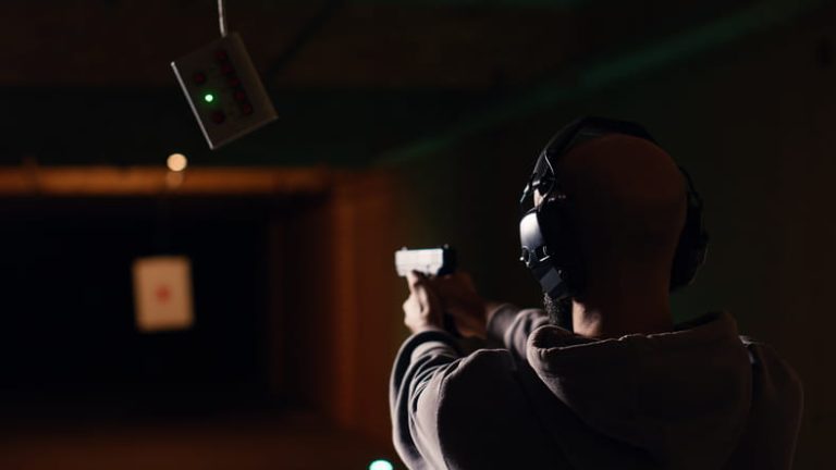 Rear view of a man wearing noise-canceling headphones aiming a handgun at a target in a dimly lit indoor shooting range.