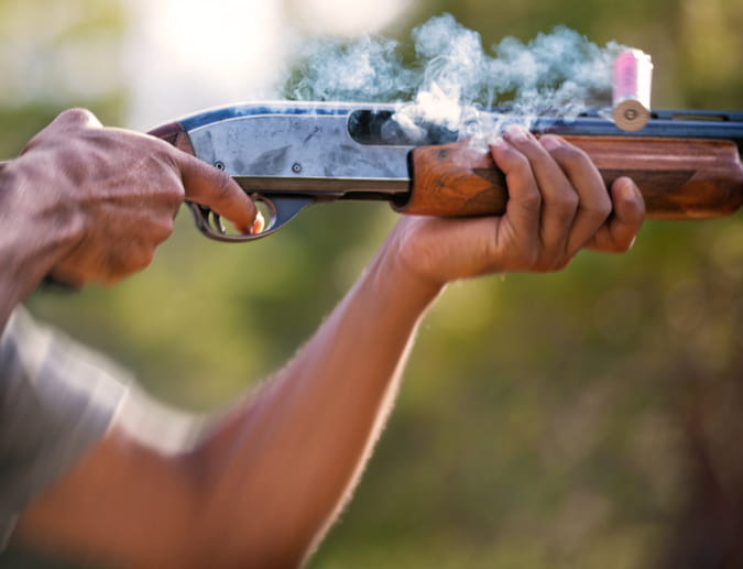 Close-up of a person firing a shotgun outdoors, capturing a smoking spent shell being ejected from the chamber against a blurred natural background.