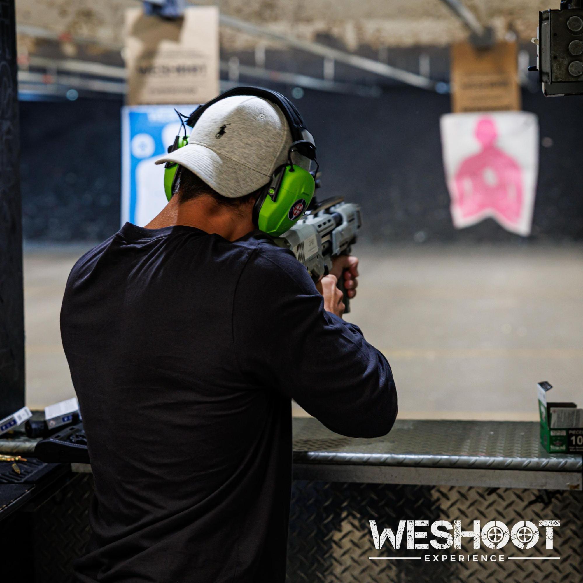A shooter wearing green ear protection and a white cap aims a firearm at a pink silhouette target at the WeShoot Experience indoor shooting range.